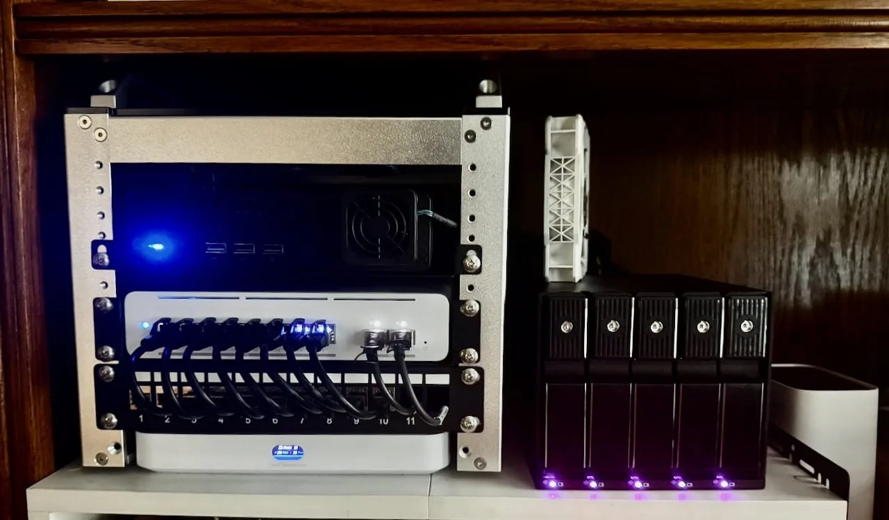 A wooden cubby holding a small open rack — mini PC on top, 10-gigabit switch and patch panel below, gateway at the bottom — next to a freestanding five-bay drive cage with status lights glowing at the base